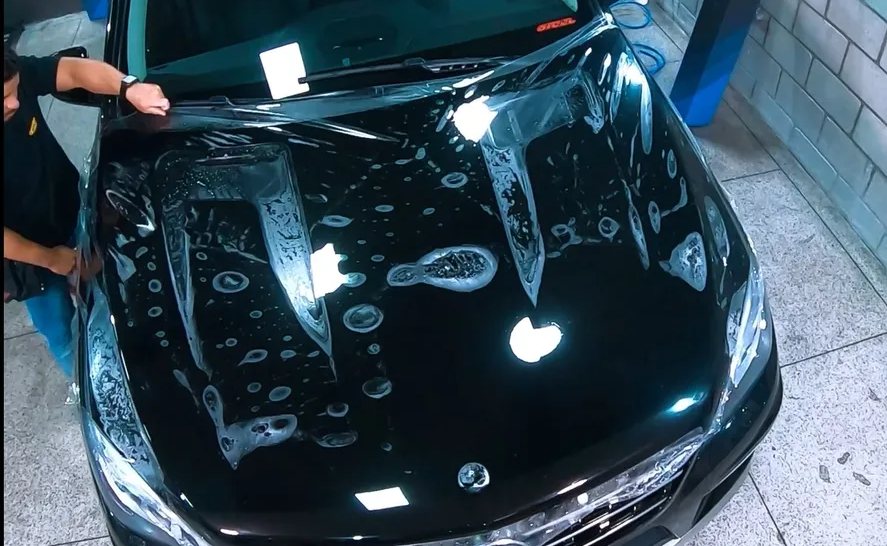 Aplicador estendendo PPF no capô de um carro preto, visto de cima